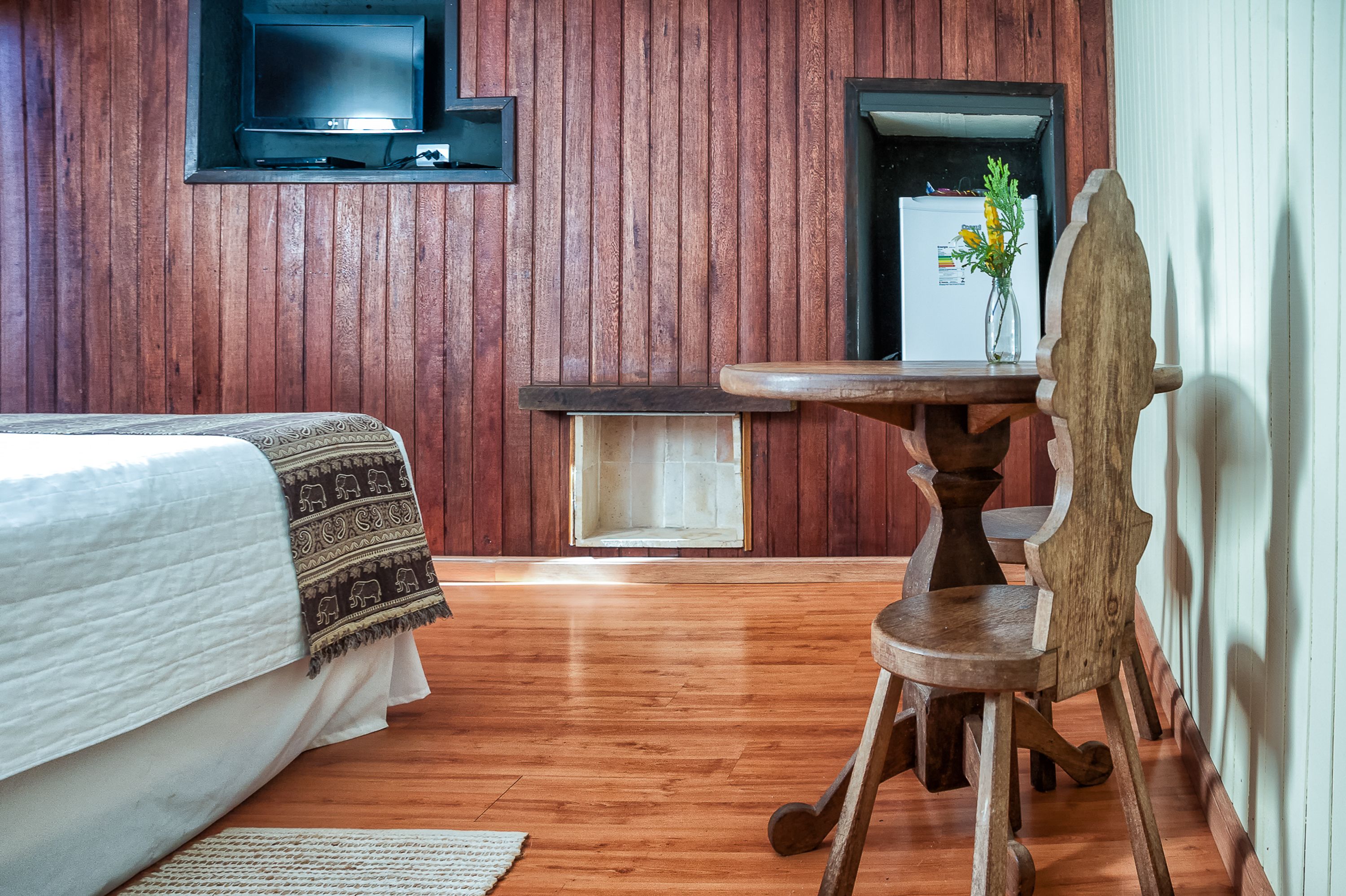 Habitación con paneles de madera, una cama, una mesita con un florero y un televisor montado en la pared.