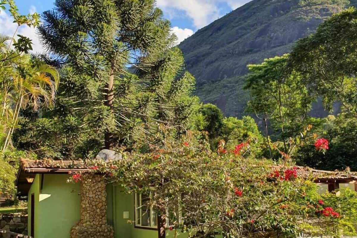 A rustic house with a stone wall, a small garden with red flowers, and a mountainous backdrop.