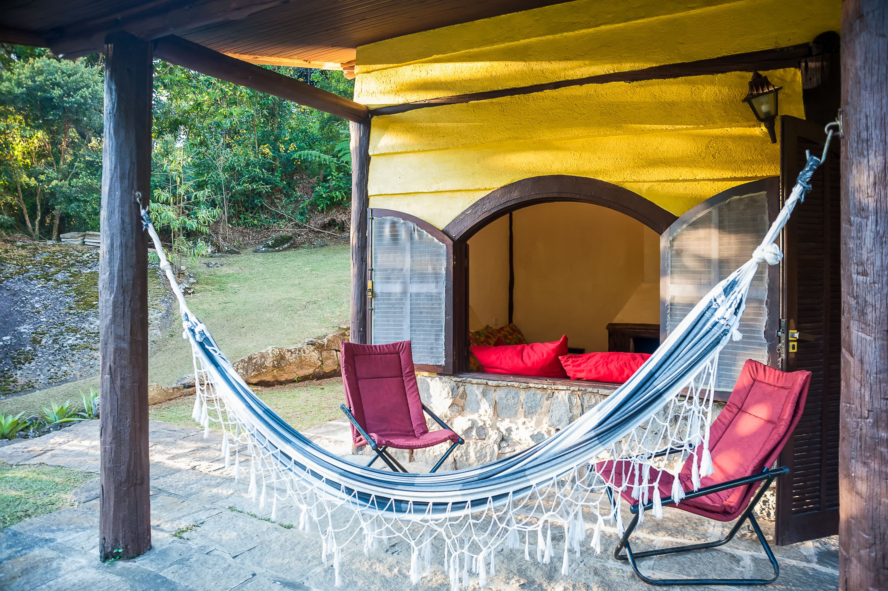 Uma cadeira de rede e uma cadeira vermelha sob um toldo amarelo, com uma parede de pedra e vista para o verde.