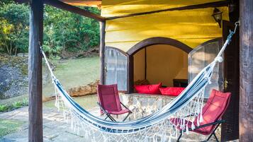 Uma cadeira de rede e uma cadeira vermelha sob um toldo amarelo, com uma parede de pedra e vista para o verde.