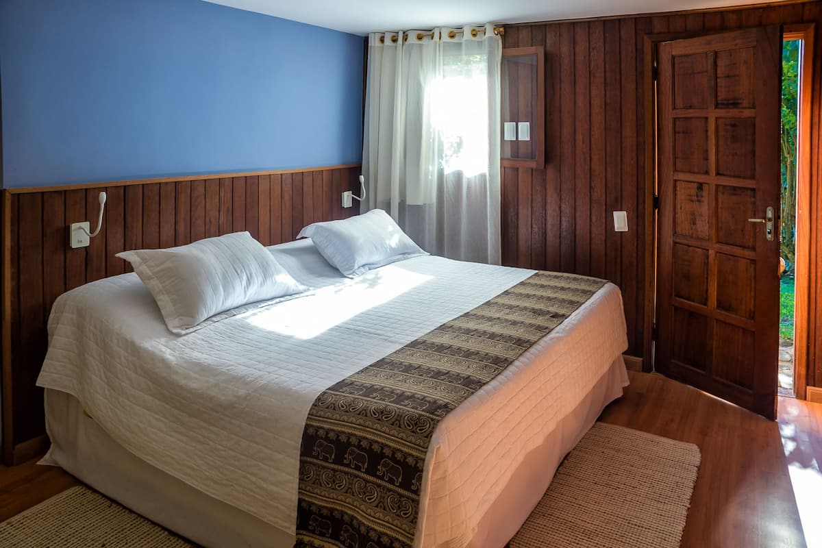 A bedroom with a large bed, wooden paneling on the walls, a window with curtains, and a door leading outside.