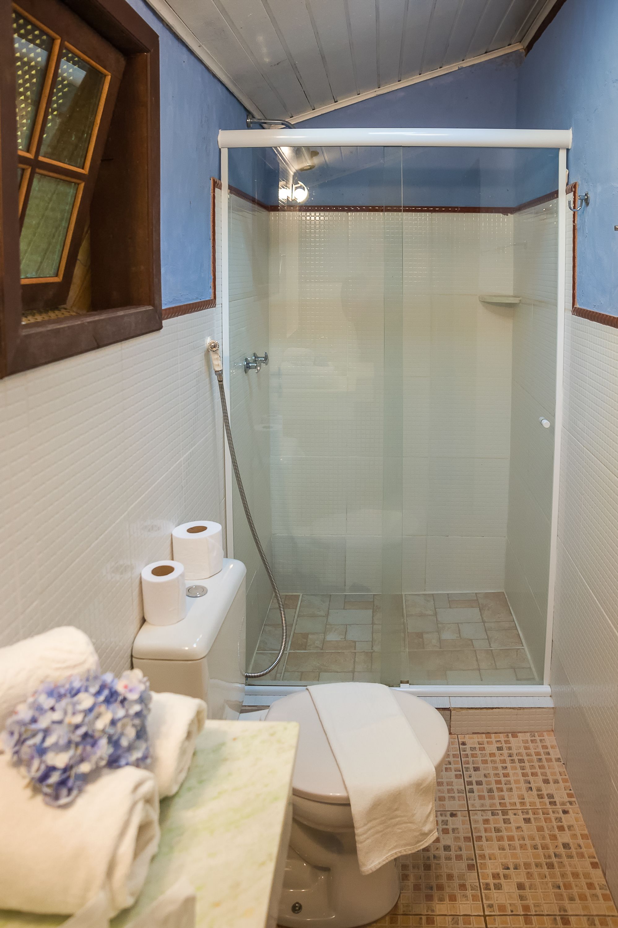 A small bathroom with a toilet, a glass-enclosed shower, and a window with blinds.