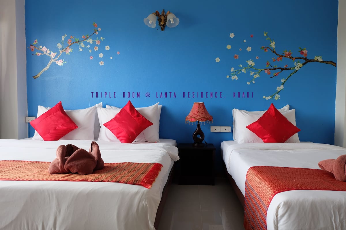 A hotel room with two beds, each with white bedding and red pillows. The room has a blue wall with a cherry blossom mural and a chandelier.