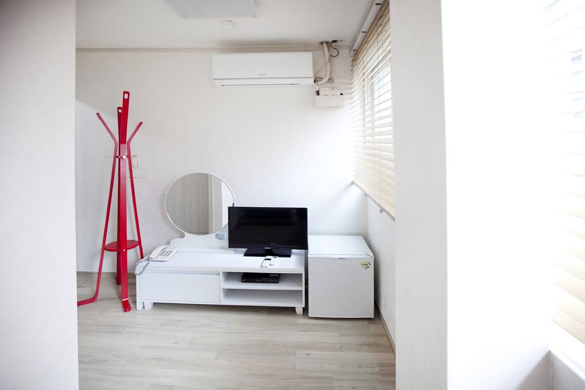A small, modern room with a white desk, a flat-screen TV, a mirror, and a red coat rack.