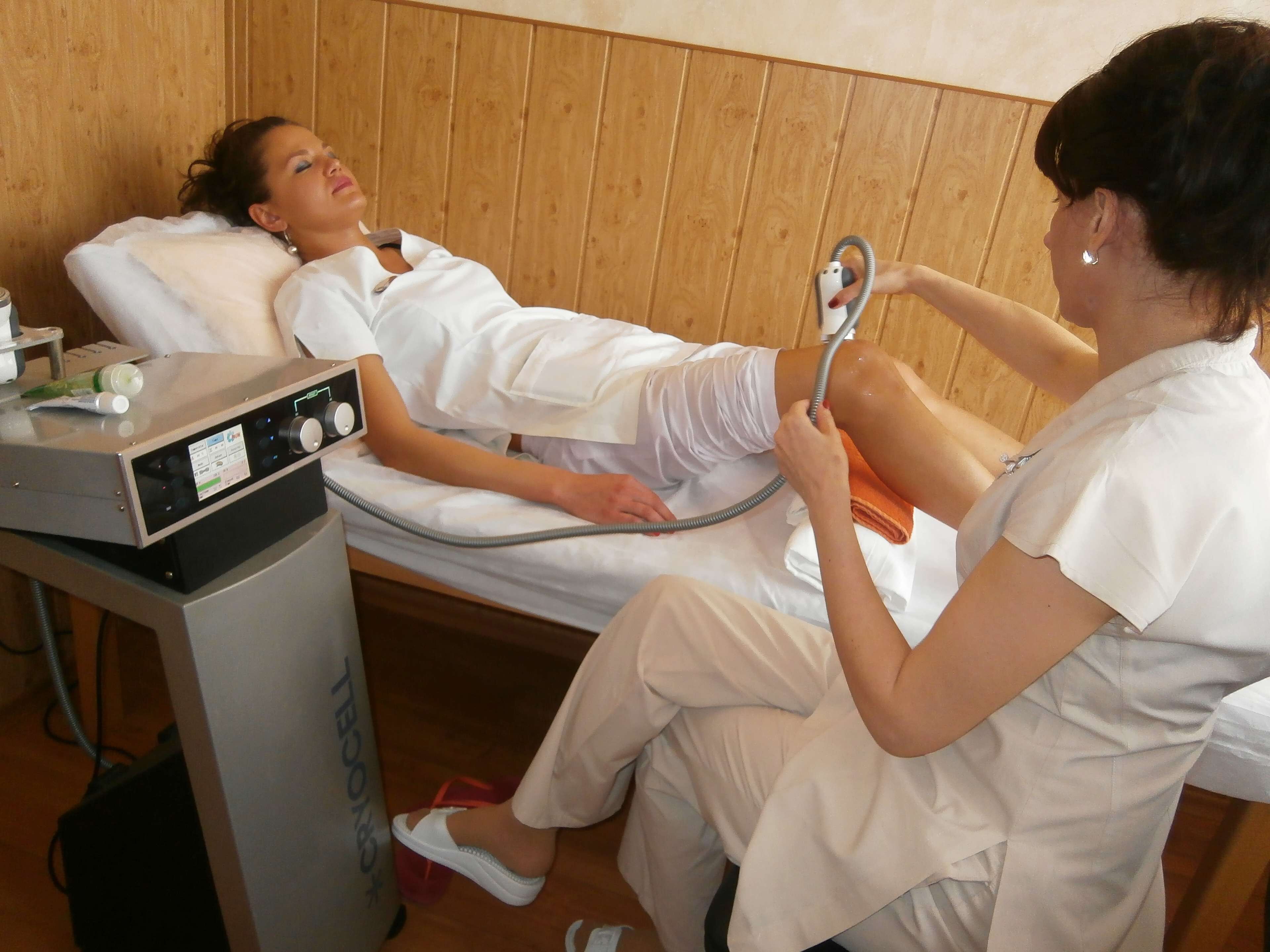 A medical examination room with a patient lying on a bed and a healthcare professional using a device on the patient's leg.