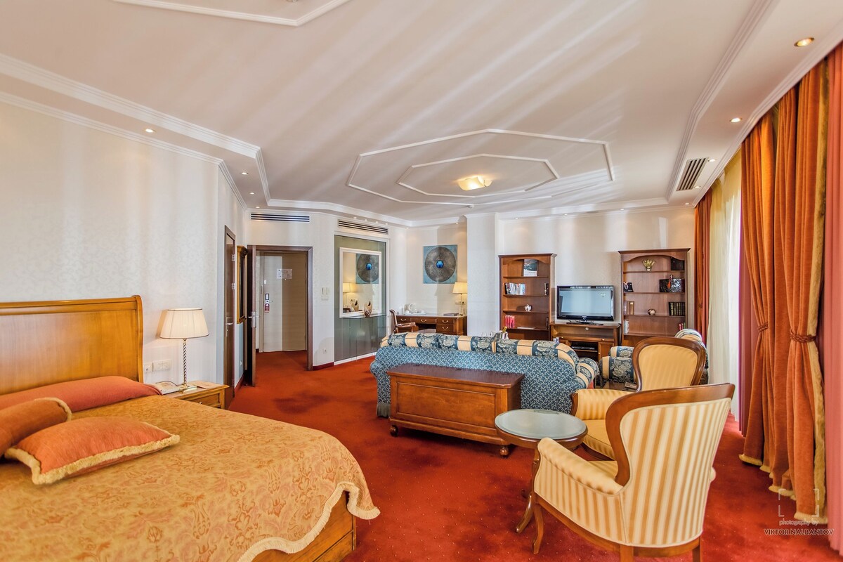 Habitación de hotel con una cama grande, un sofá, una mesita y un televisor. Se ve una puerta que da a un baño y un pasillo.