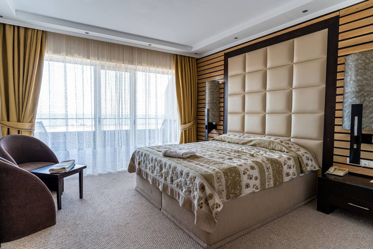 Un dormitorio con una cama grande, una mesita de noche, una silla, una ventana con cortinas y un espejo montado en la pared.