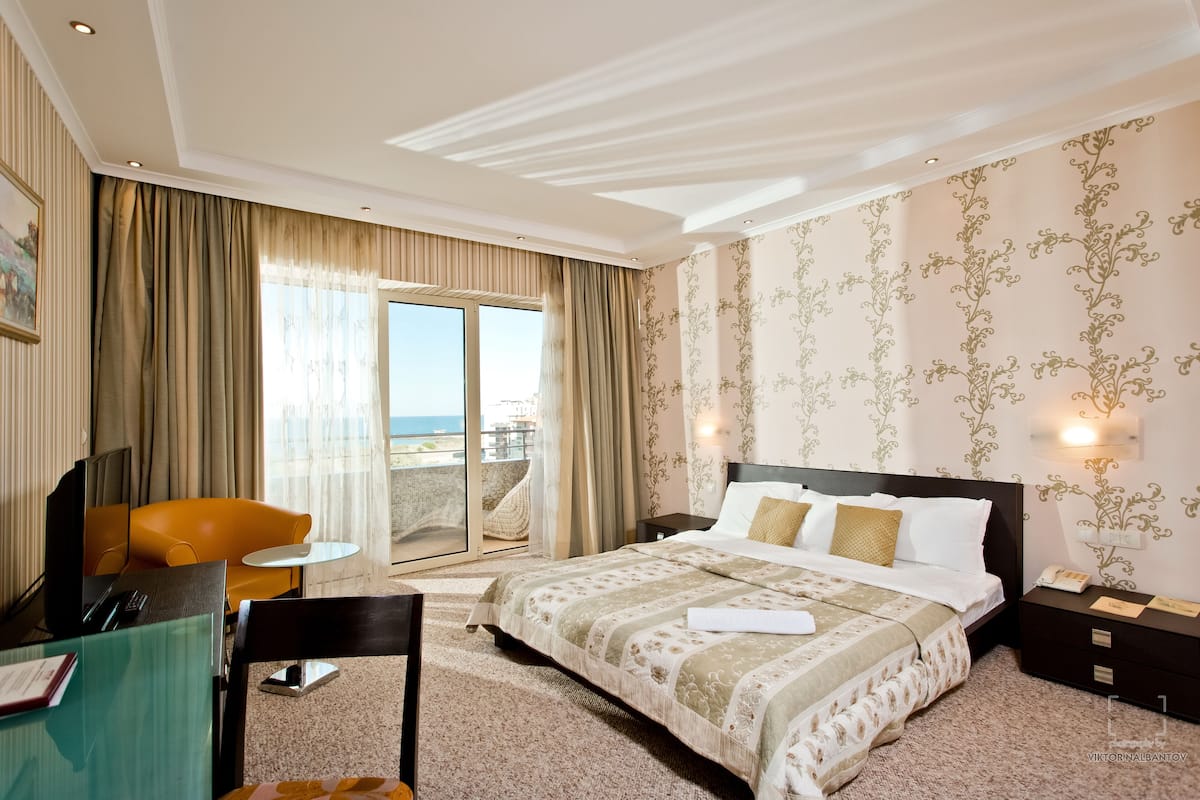 Habitación de hotel con una cama grande, un escritorio con silla, una mesita y vista al mar a través de una puerta corredera.