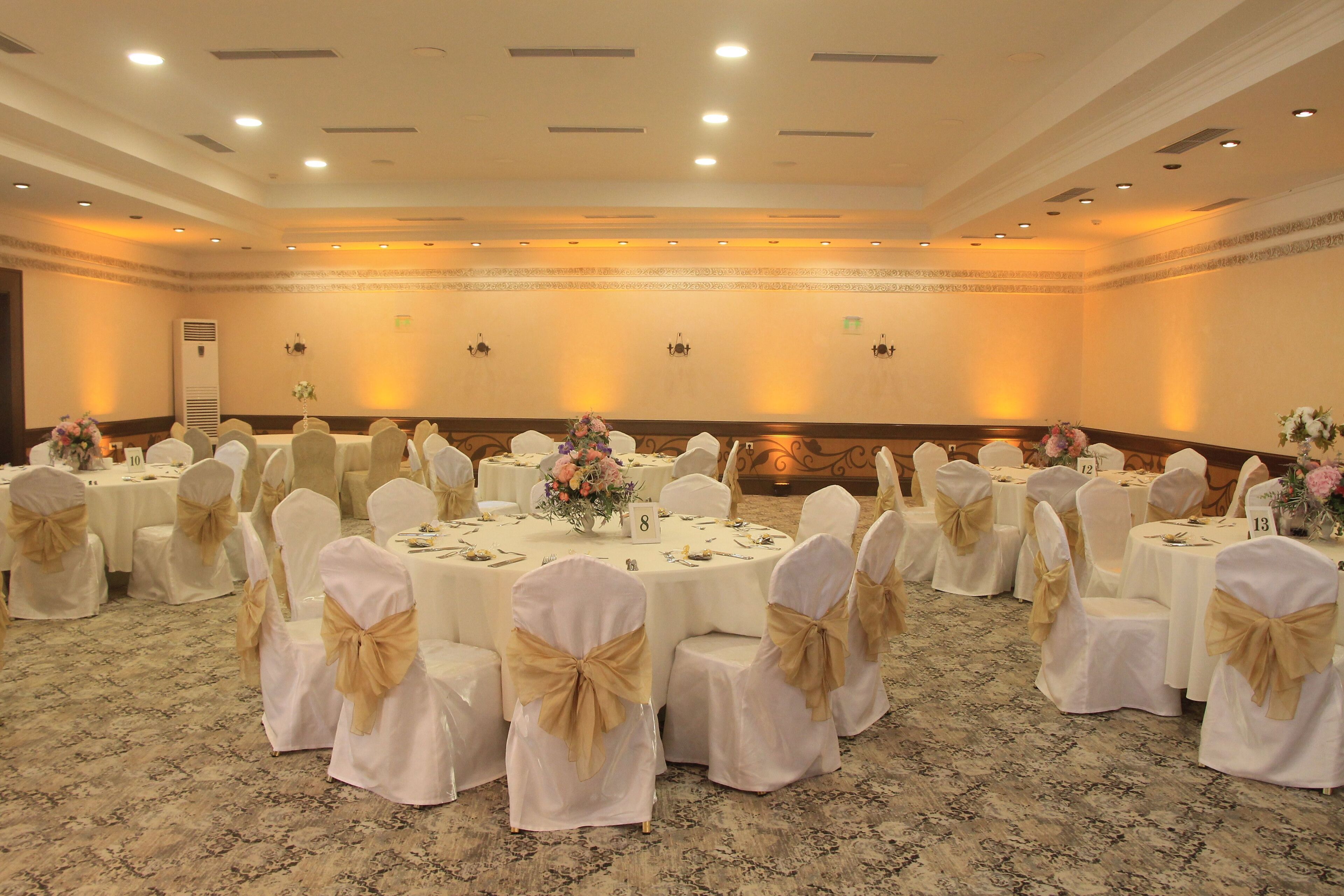 A banquet hall with round tables, white chairs with gold sashes, and floral centerpieces.