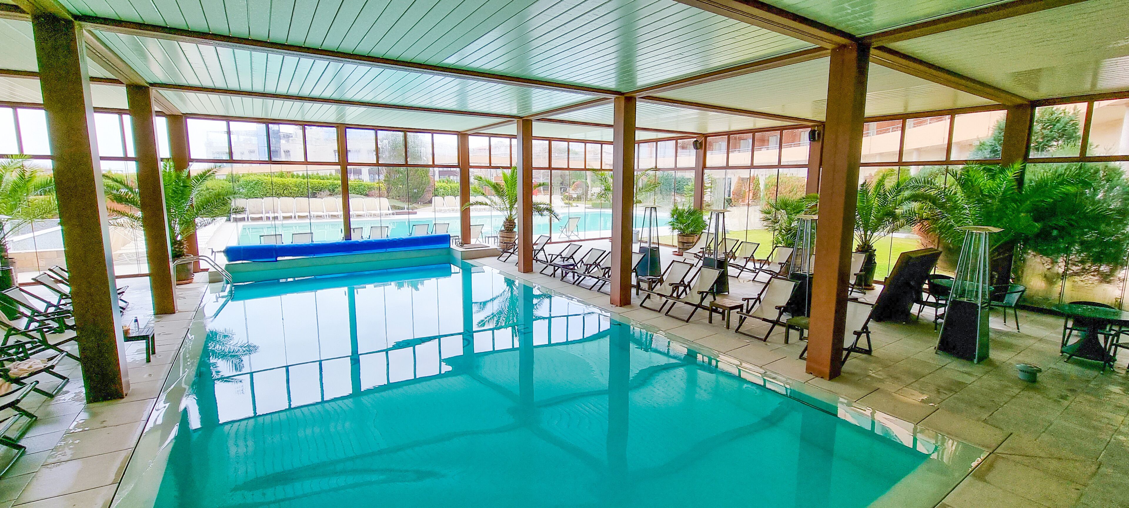 An indoor swimming pool with clear glass walls, surrounded by lounge chairs and potted plants.