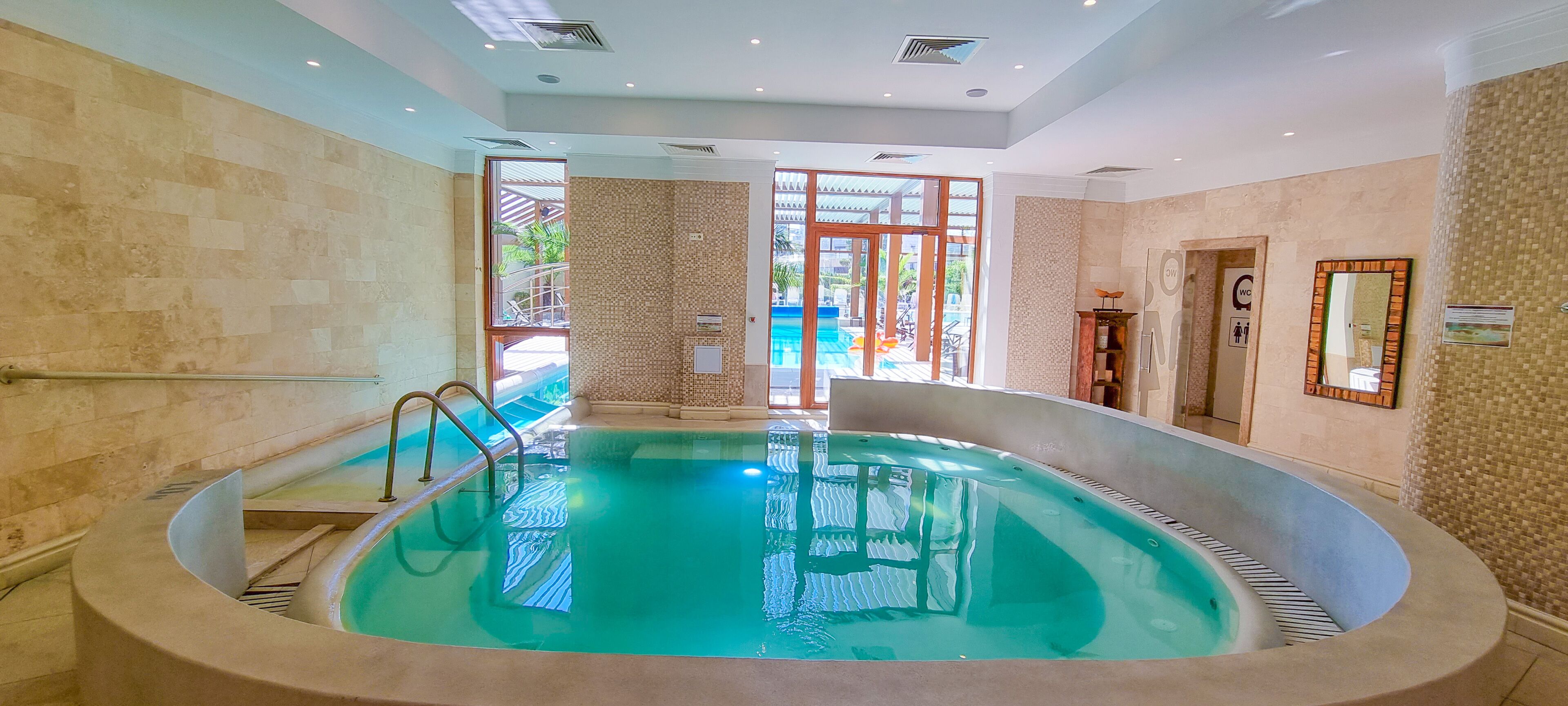 Una amplia piscina interior con pared de cristal, una escalera metálica y una puerta que da al exterior.