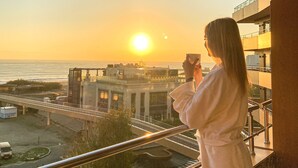 Una persona fotografiando la puesta de sol desde un balcón con vistas a un edificio moderno y una calle con vehículos.