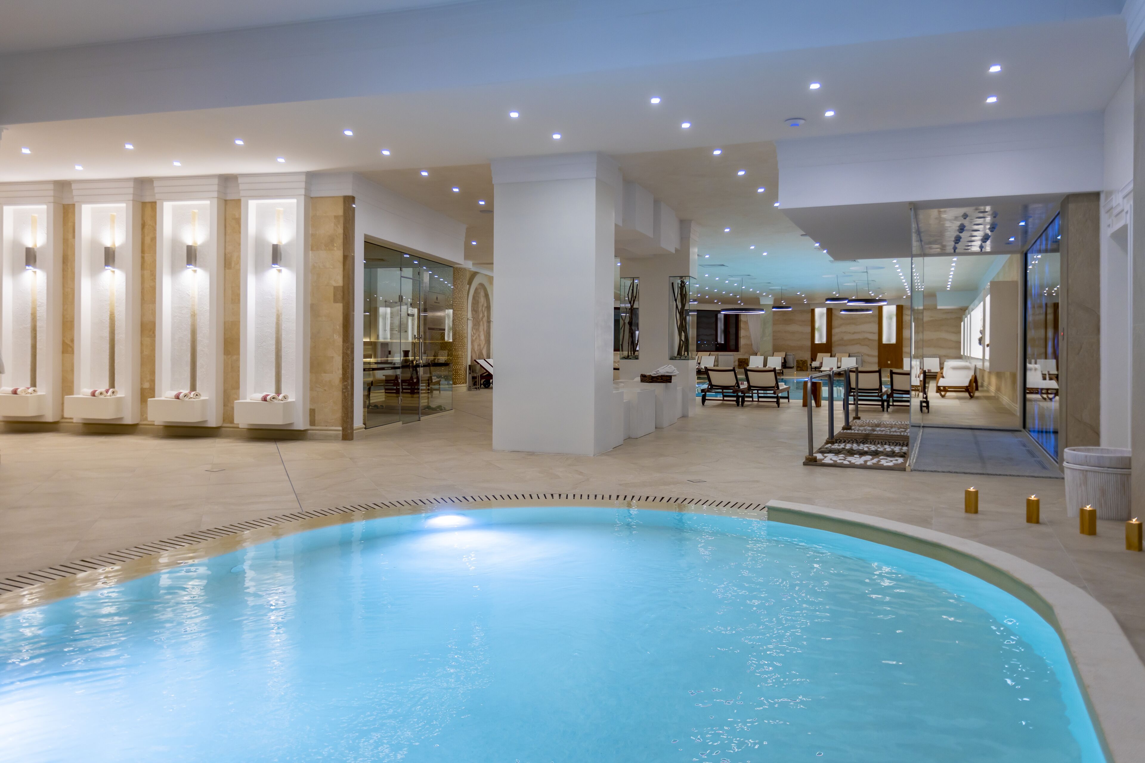 A modern indoor pool area with a clear pool, seating areas, and a reception desk.