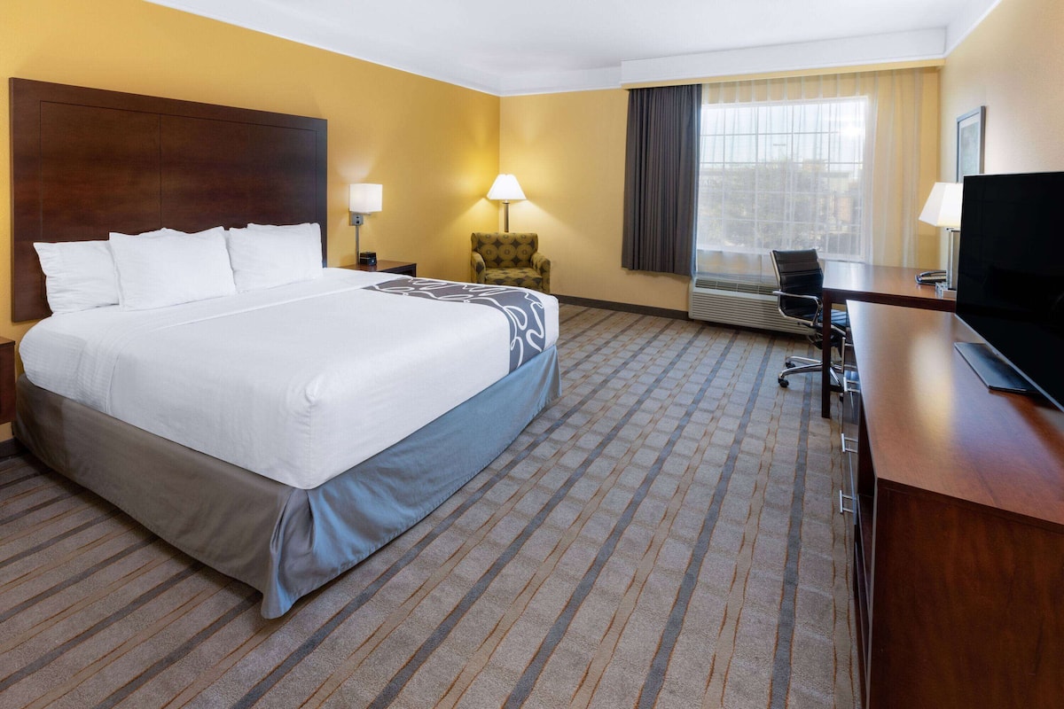 A hotel room with a large bed, a desk with a computer, a chair, a lamp, and a window with curtains.