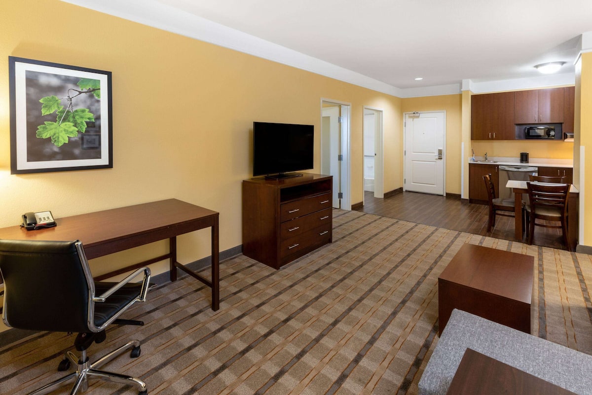A hotel room with a desk, chair, television, kitchenette, and a framed picture on the wall.