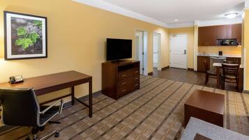 Ein Hotelzimmer mit Schreibtisch, Stuhl, Fernseher, Küchenzeile und einem gerahmten Bild an der Wand.