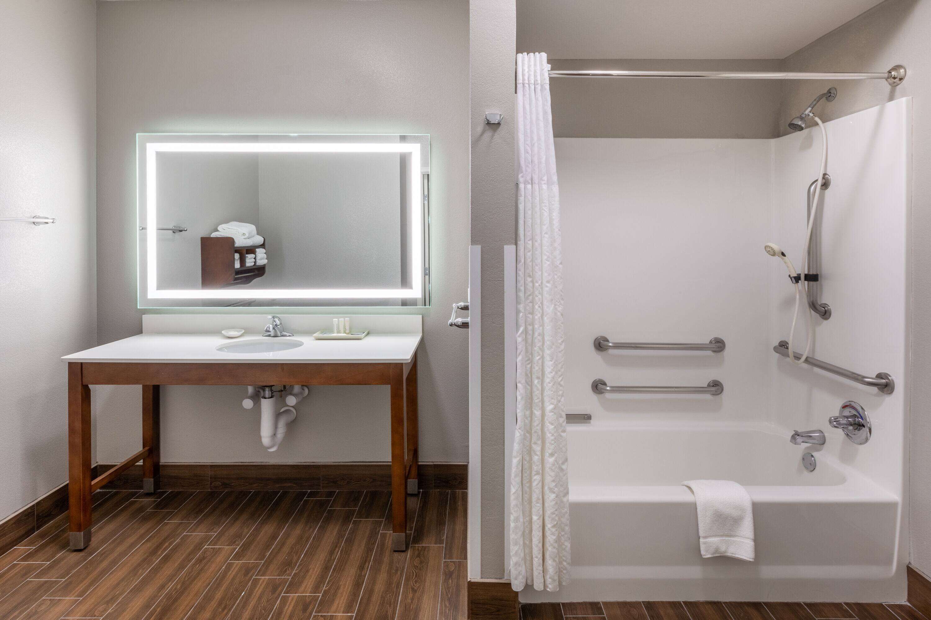 A bathroom with a wooden vanity, a large mirror with lights, a bathtub, and a shower with grab bars.