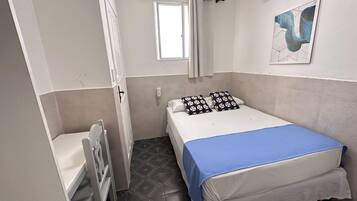A small, neatly arranged bedroom with a single bed, a bedside table, a chair, a window, and a door.