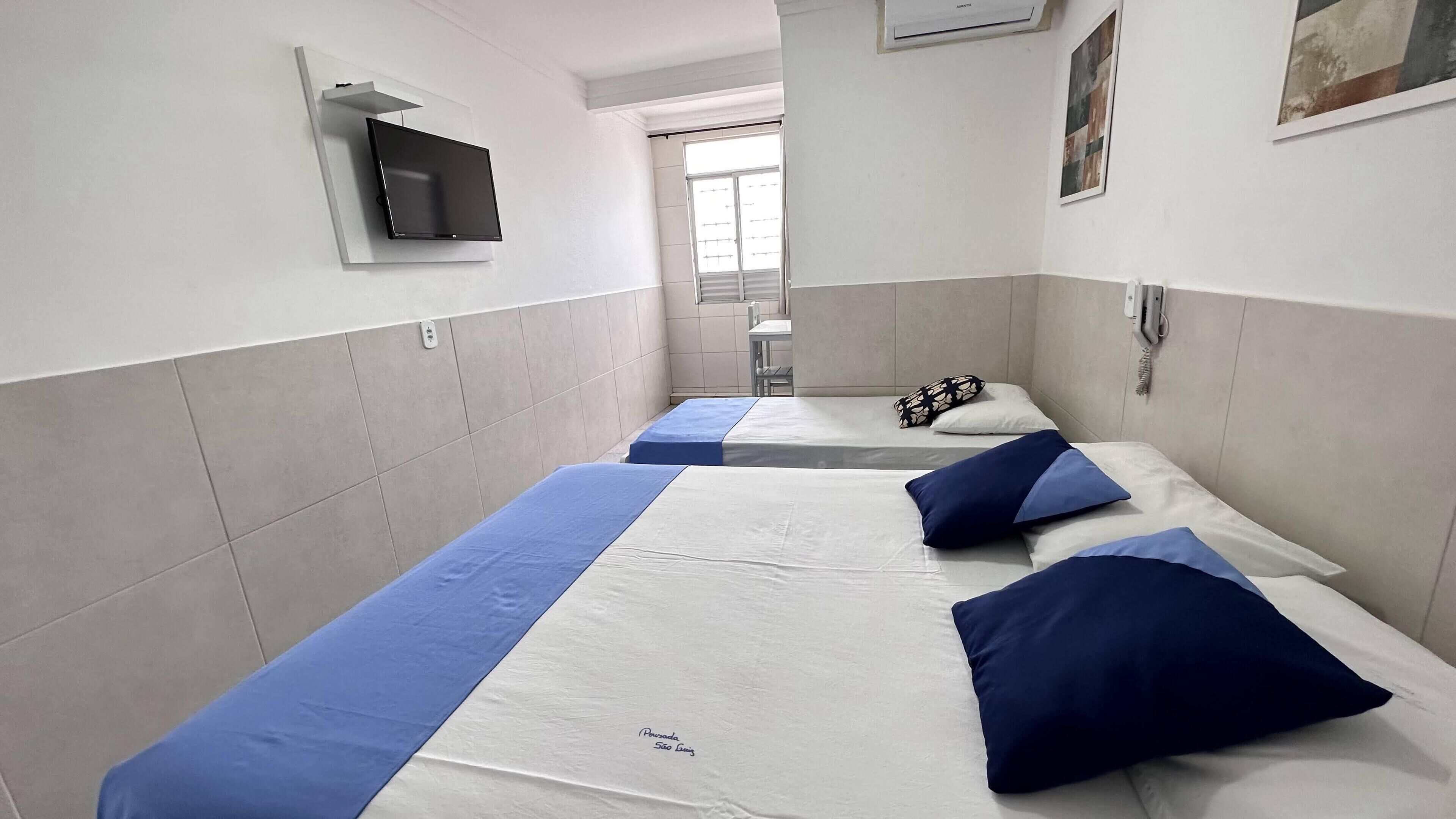 Quarto de hotel com três camas, cada uma com colcha azul e branca. Há uma televisão de tela plana fixada na parede acima das camas.