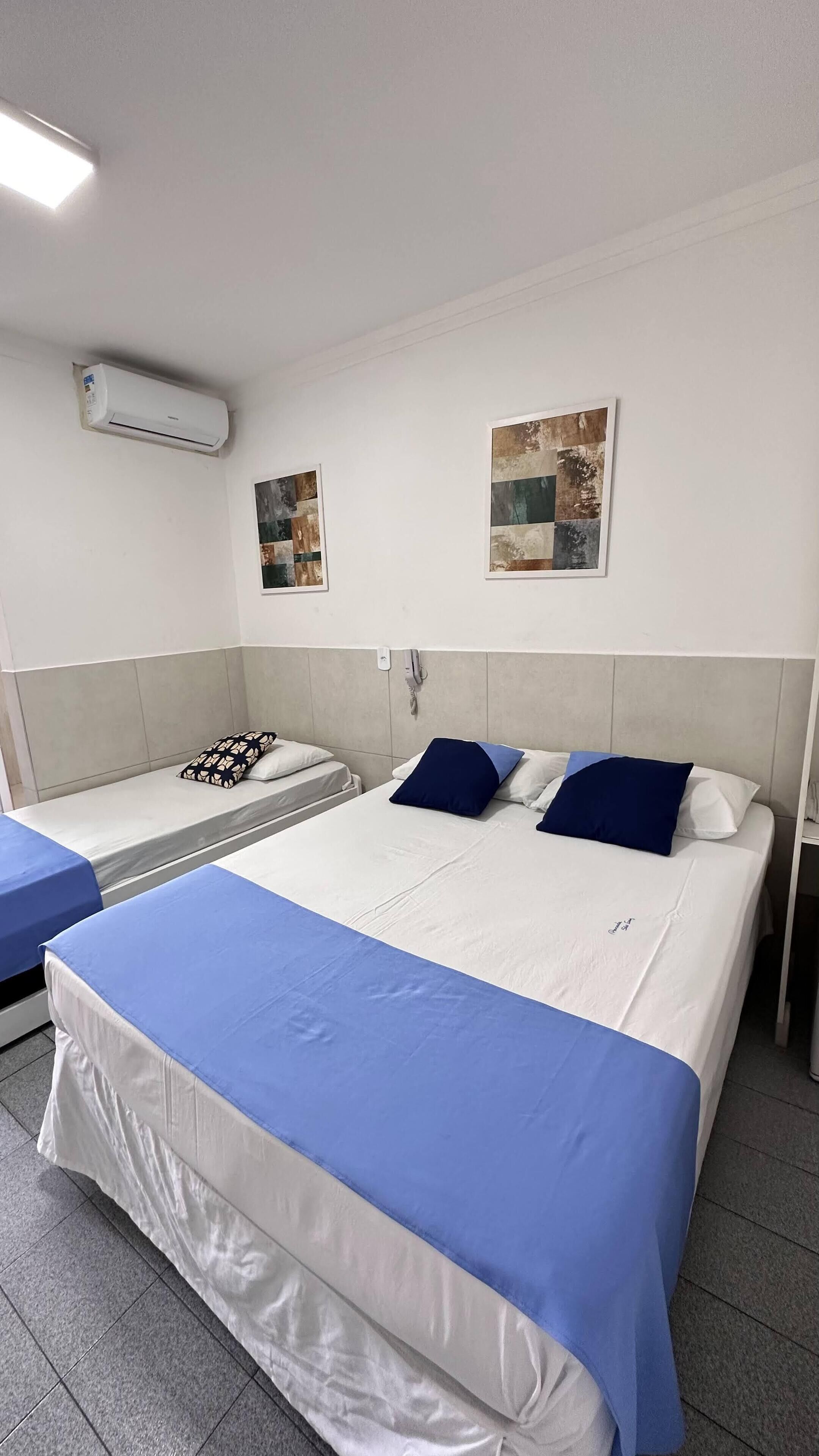 Habitación de hotel con una cama grande, dos mesitas de noche, un equipo de aire acondicionado y dos obras de arte colgadas en la pared.