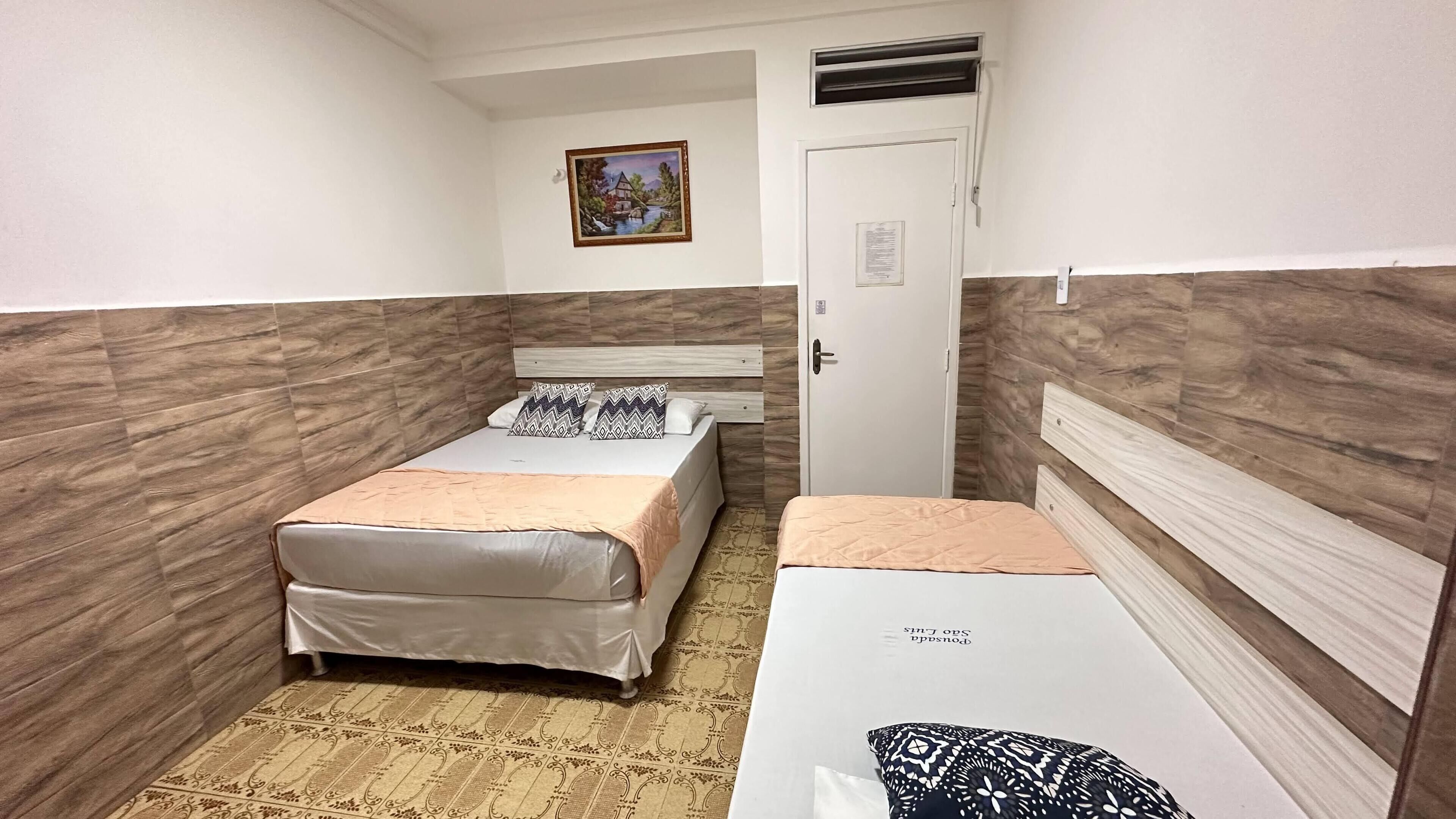 Una habitación pequeña y estrecha con dos camas, una con cabecera y la otra sin. Se observa una puerta con un aviso, un cuadro pequeño en la pared y una alfombra estampada en el suelo.