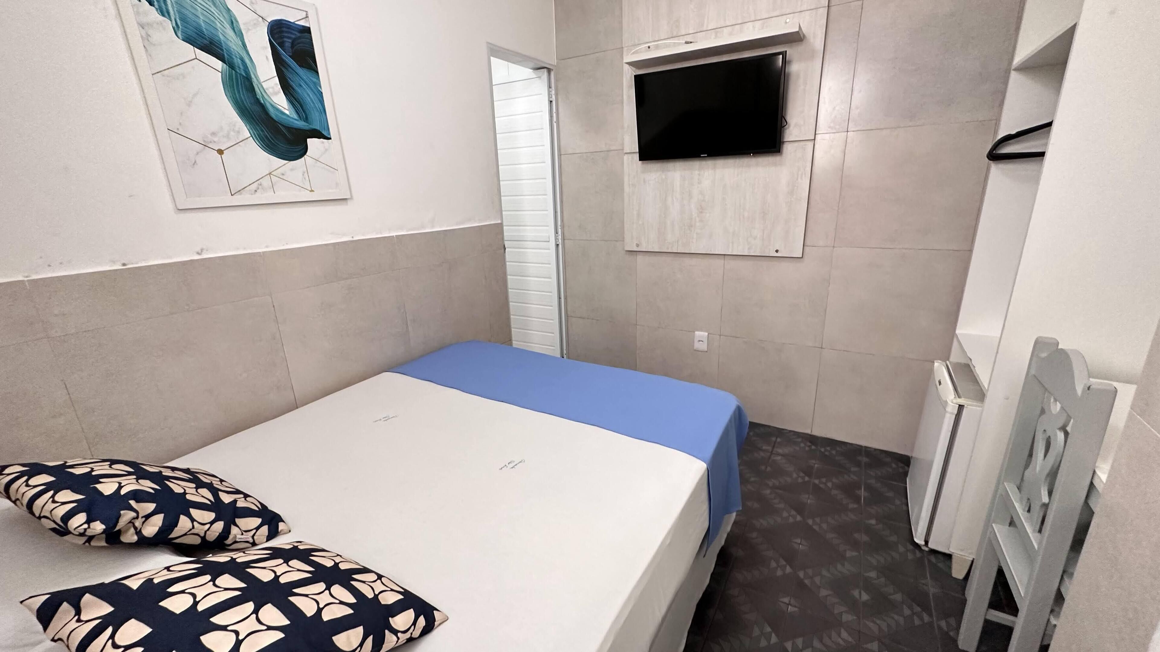 Una habitación pequeña y ordenada con una cama individual, un televisor montado en la pared y una puerta que da a otra habitación.