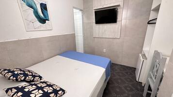 Una habitación pequeña y ordenada con una cama individual, un televisor montado en la pared y una puerta que da a otra habitación.