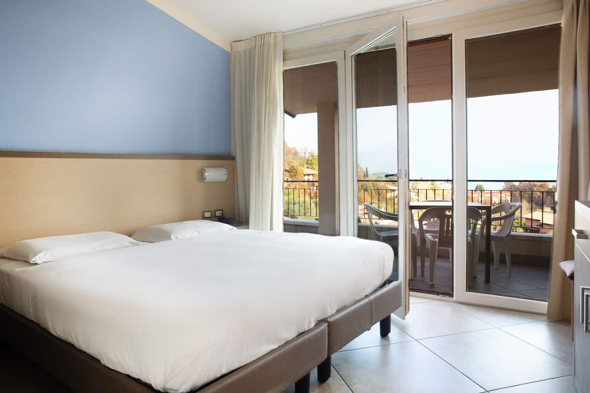 A hotel room with a large bed, a balcony with chairs, and a view of the outdoors.