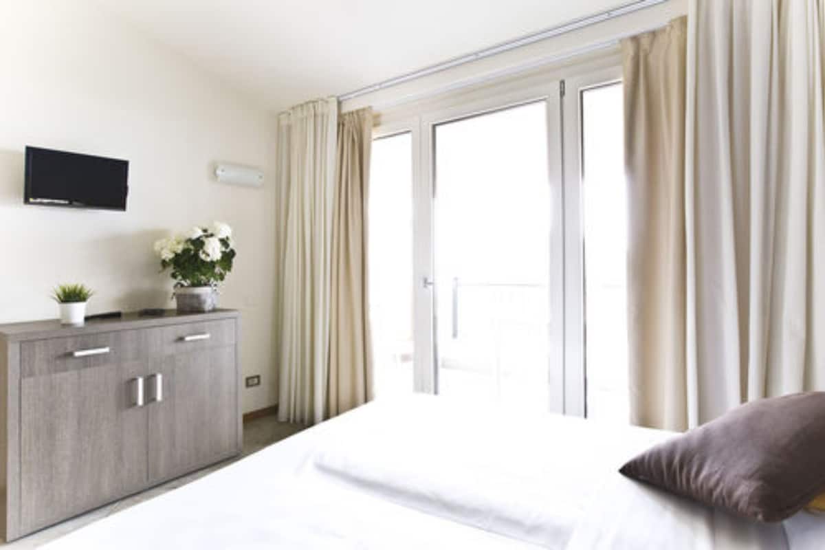 A hotel room with a bed, a TV mounted on the wall, a cabinet with a plant, and a sliding glass door to the outside.