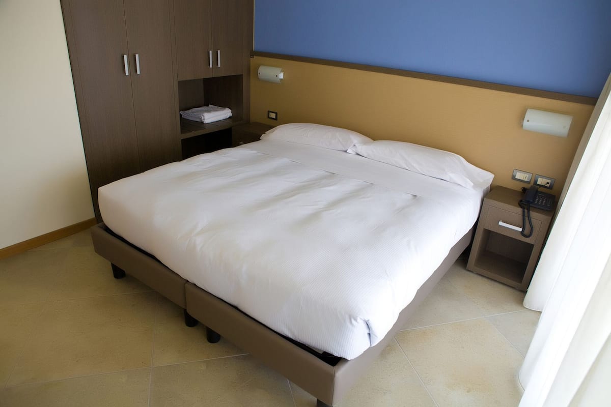 A neatly made bed in a hotel room with a wooden wardrobe, a bedside table with a phone, and a wall-mounted light fixture.