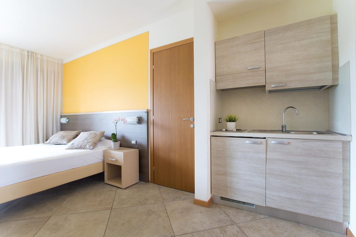A modern hotel room with a bed, bedside table, and a small kitchen area including a sink and cabinets.