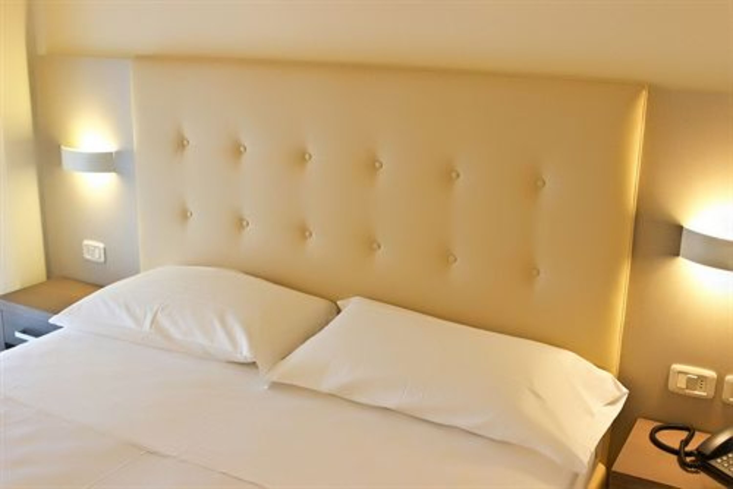 Et hotelværelse med en betrukken sengegavl, to hvide puder, et natbord med lampe og telefon.
