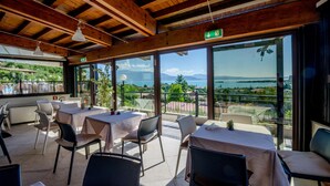Un área de comedor al aire libre con mesas y sillas, amplias ventanas que ofrecen vistas a un lago y montañas, y vigas de madera en el techo.