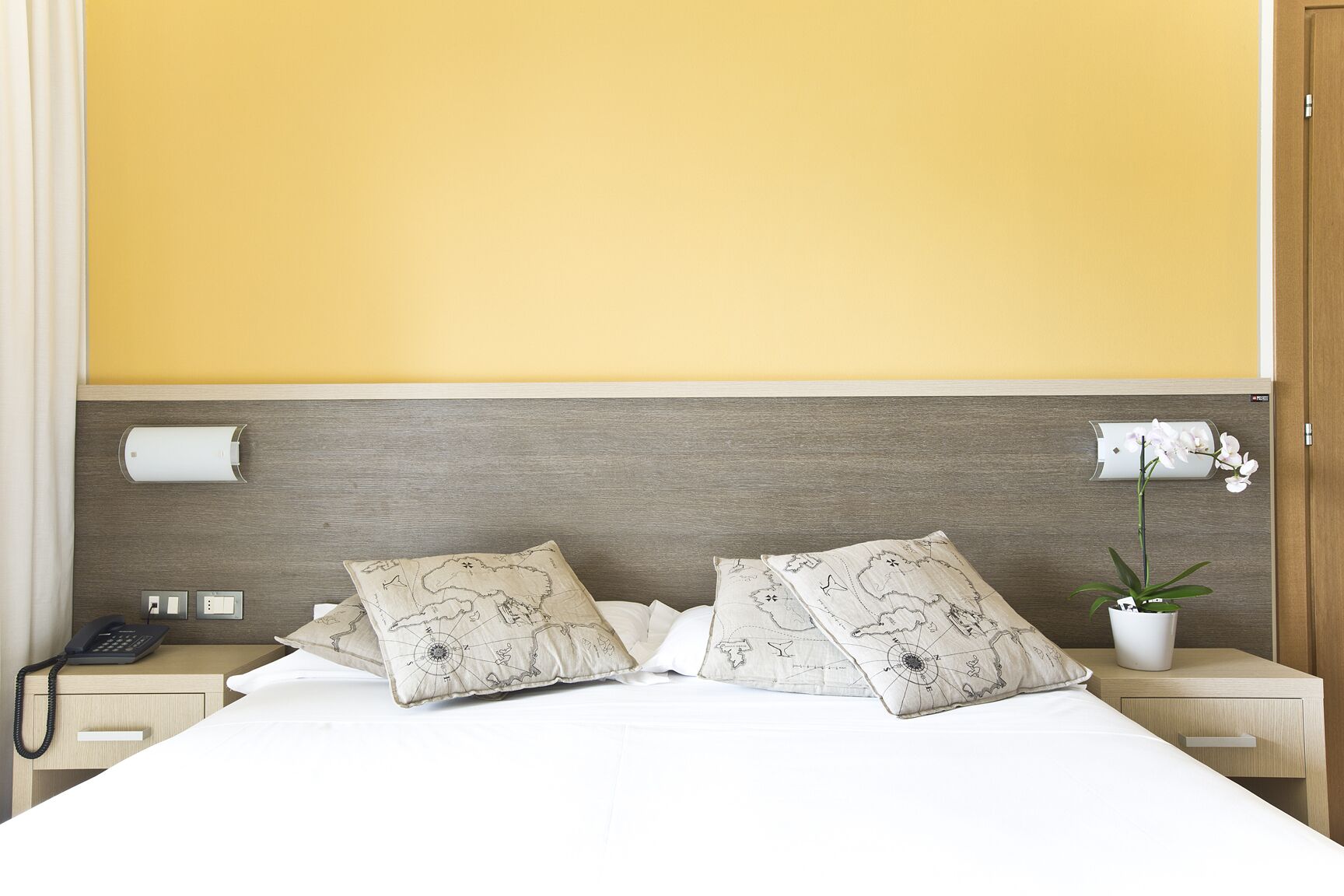 Habitación de hotel con una cama grande, dos almohadas con diseño de mapa, un teléfono en la mesita de noche, una mesita de noche, una lámpara de pared y una orquídea en maceta.