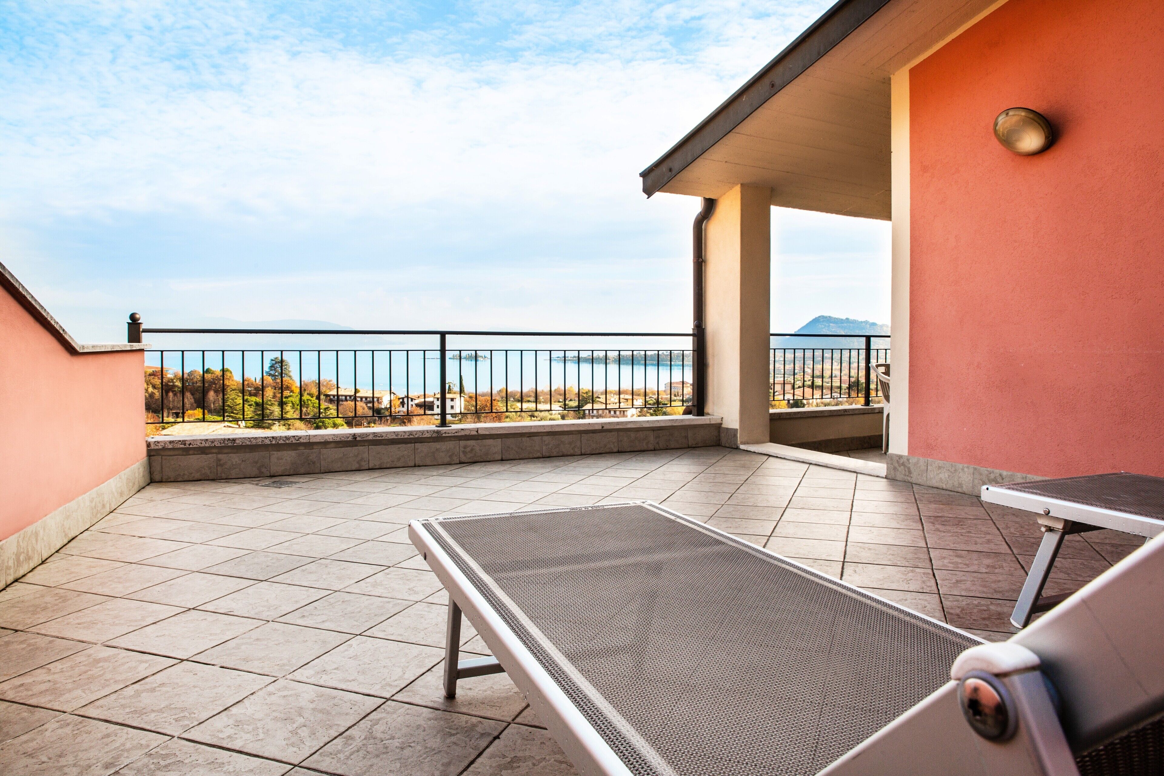 Un balcón con barandilla metálica, una silla de descanso y vistas al mar y montañas lejanas.
