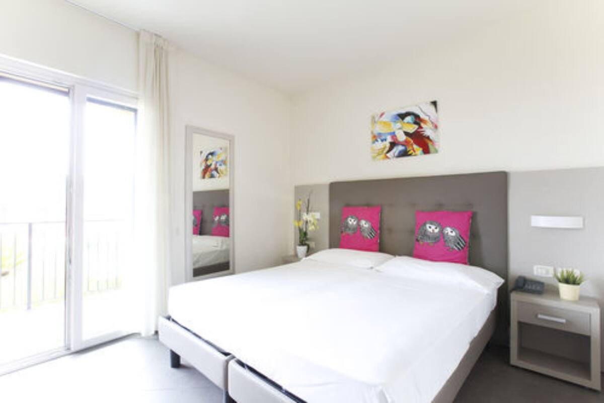 A modern hotel room with a large bed, a bedside table with a lamp, a small bedside plant, and a window with curtains.