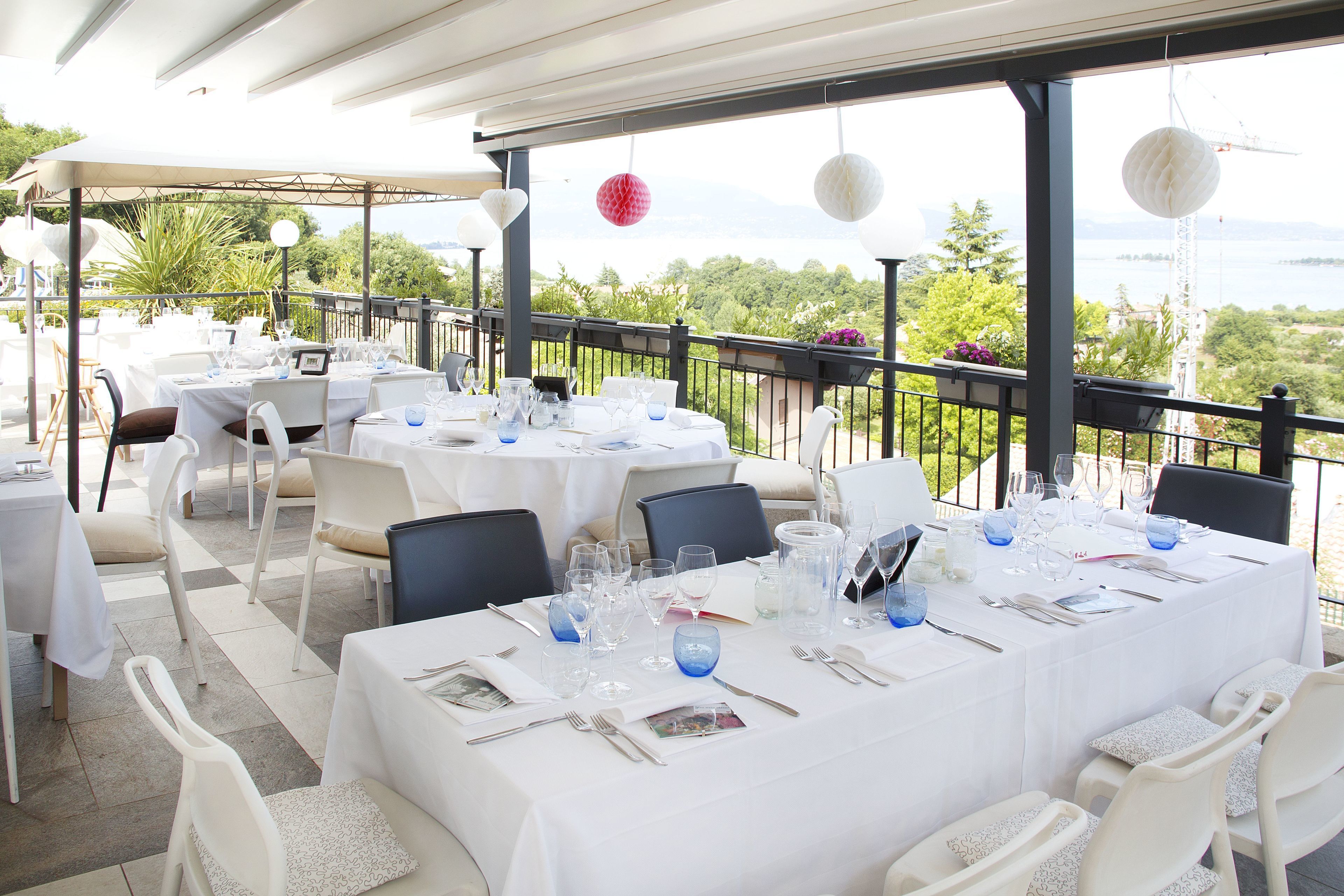 An outdoor dining area with tables set for two, white chairs, and a view of the sea.