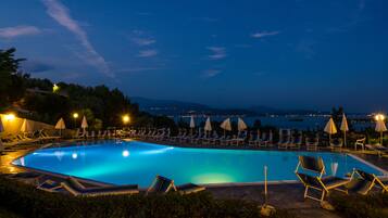 A swimming pool with lounge chairs and umbrellas, surrounded by greenery and overlooking a body of water with distant city lights.