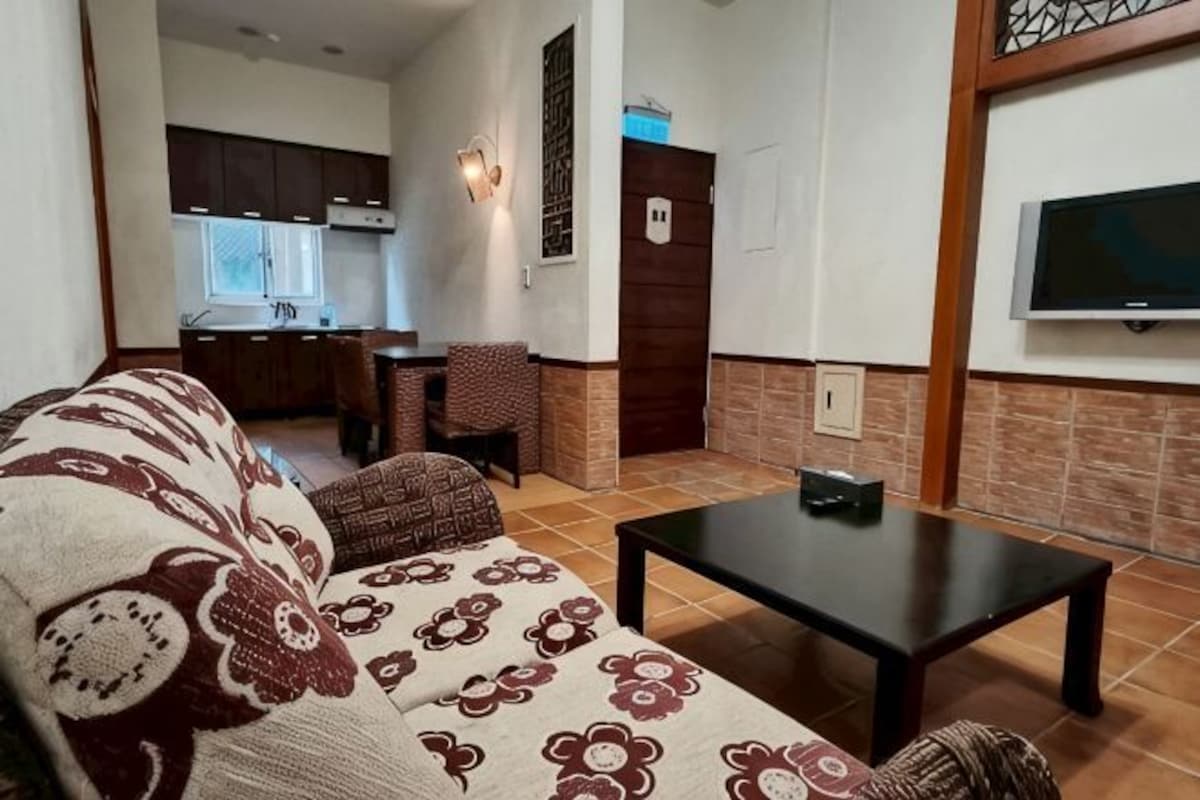 A living room with a sofa, a coffee table, a TV, and a kitchen in the background.
