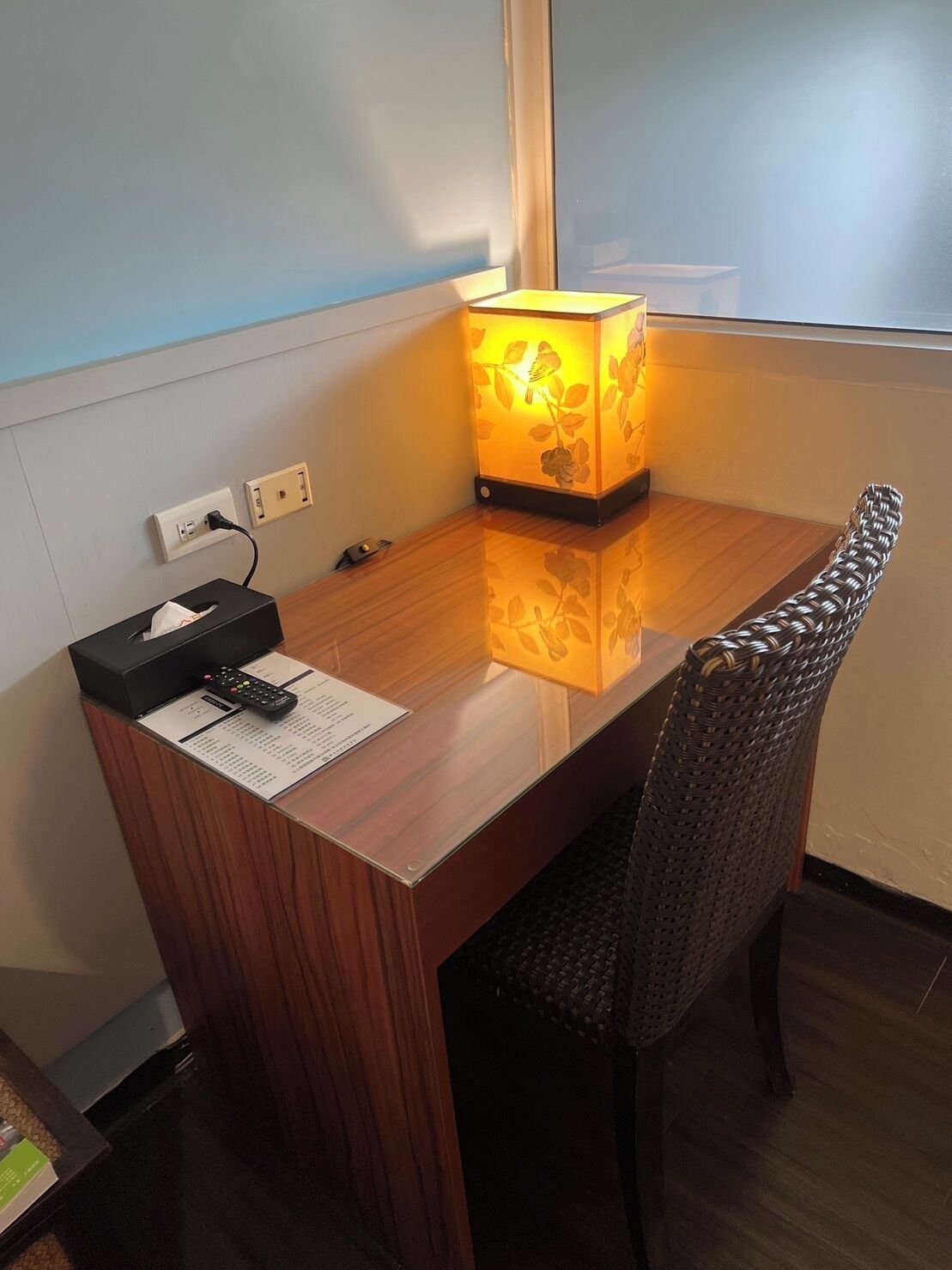 A wooden desk with a lamp, a tissue box, a remote control, and a menu on a table.