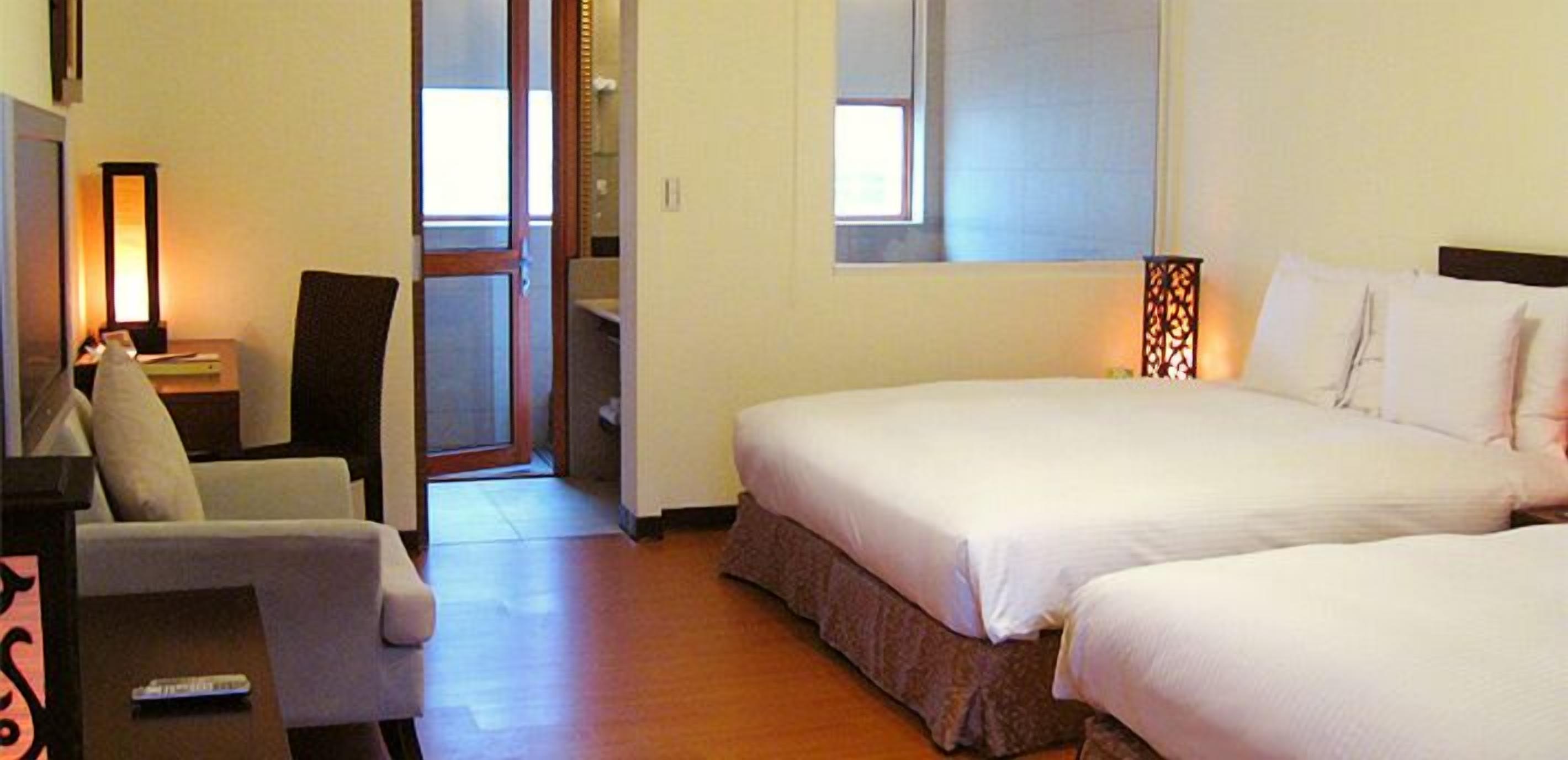 A hotel room with two beds, a chair, a desk with a lamp, and a window.