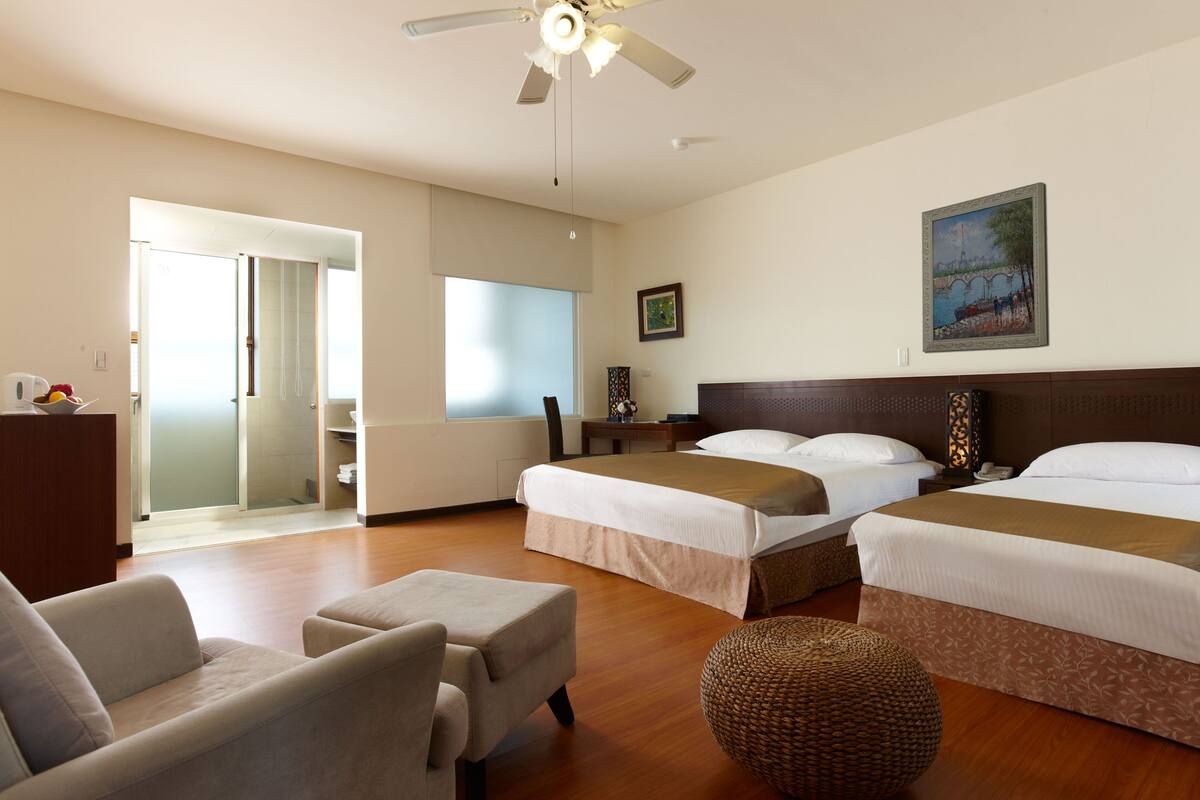 Quarto de hotel com duas camas, um sofá, uma poltrona, uma mesinha e um ventilador de teto.