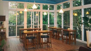 Ein Wintergarten mit Holztischen und -stühlen, großen Fenstern und hängenden Pendelleuchten.