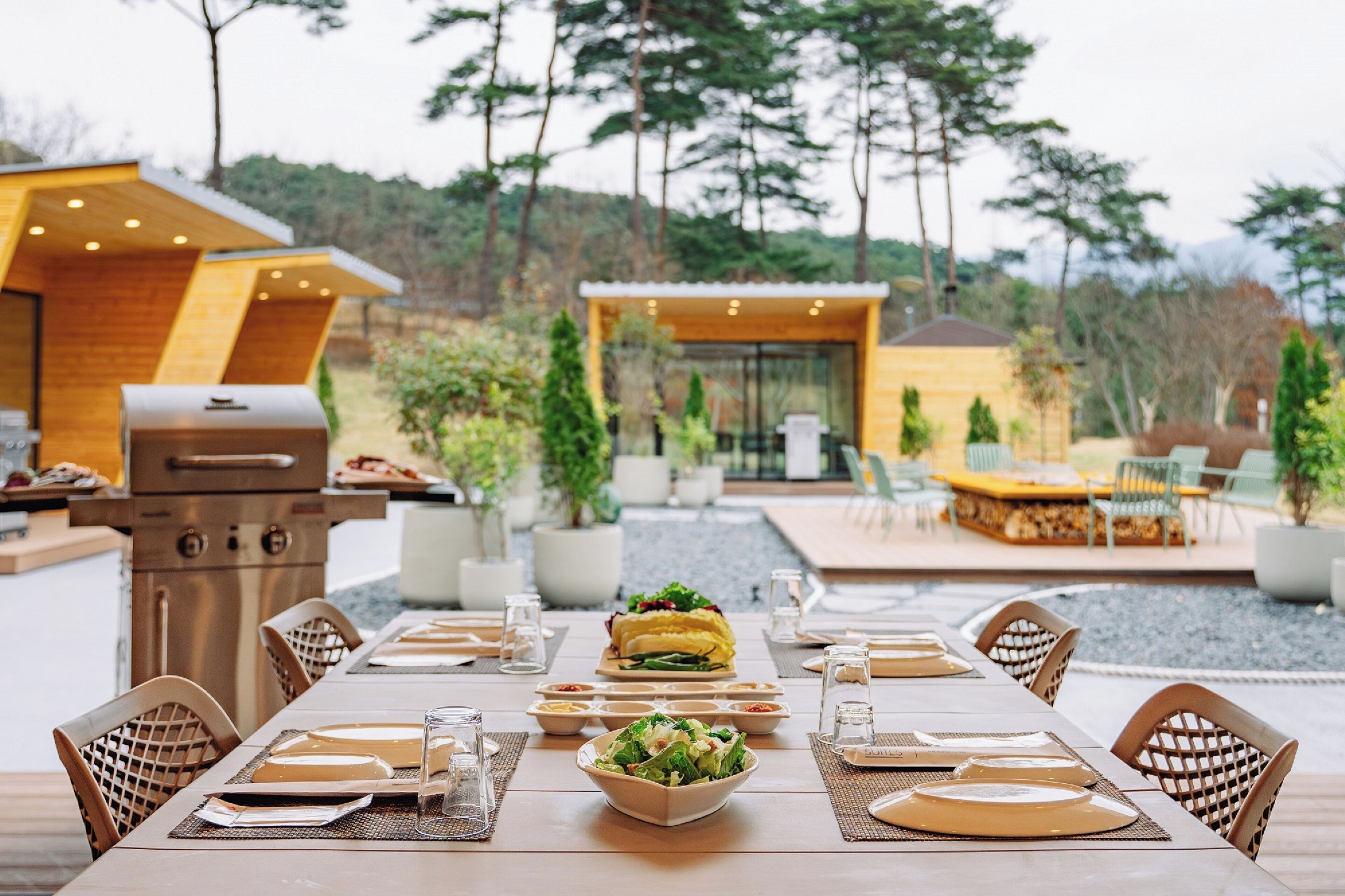 An outdoor dining area with a long table set for a meal, surrounded by chairs. There is a grill and a small structure with a glass top in the background.