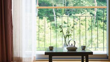 A table with a vase of greenery and two cups, positioned by a window with a view of a balcony and greenery outside.