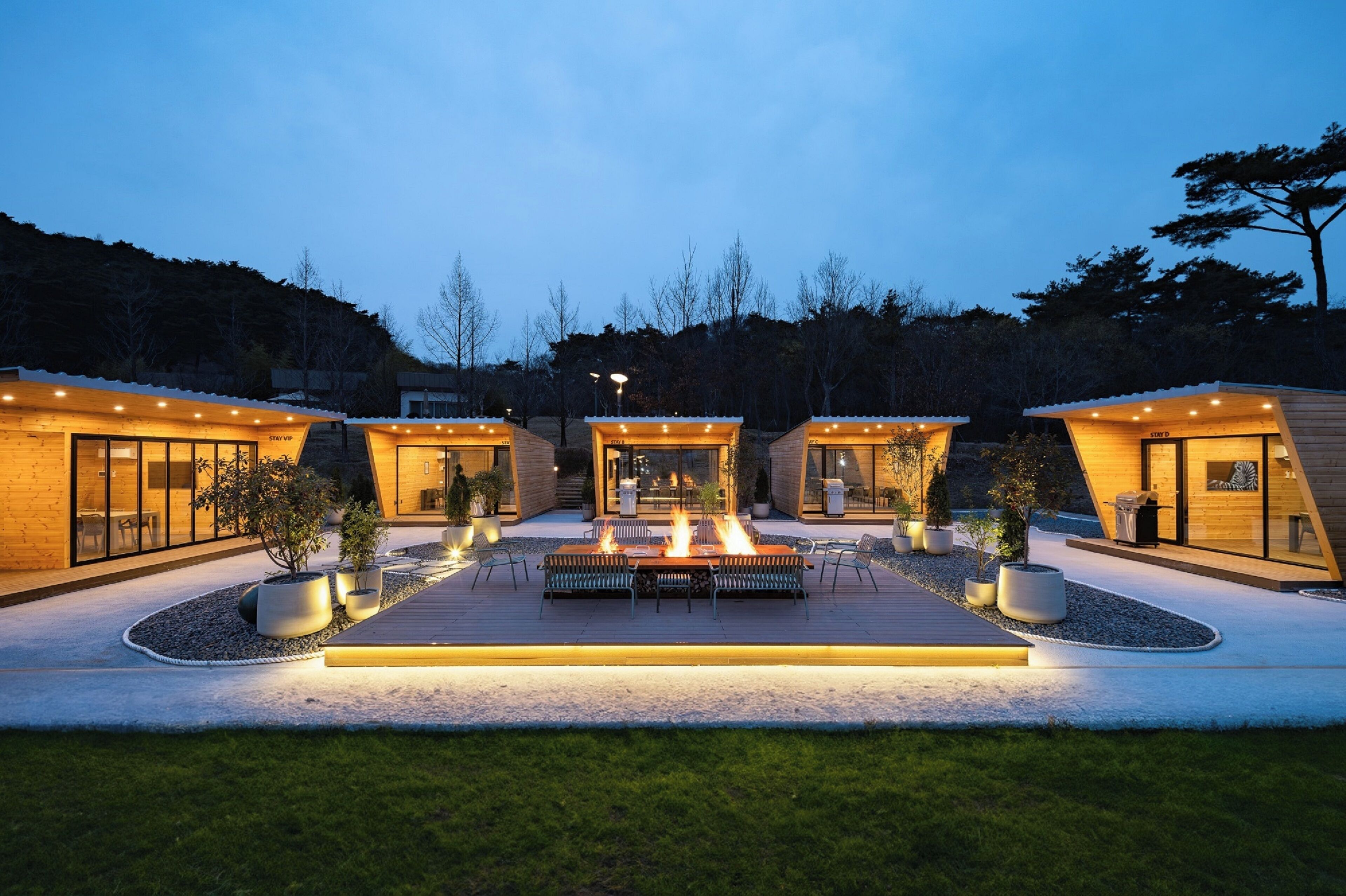 A modern outdoor patio area with a central fire feature, surrounded by three wooden cabins with glass doors and windows, and a well-maintained garden with potted plants.