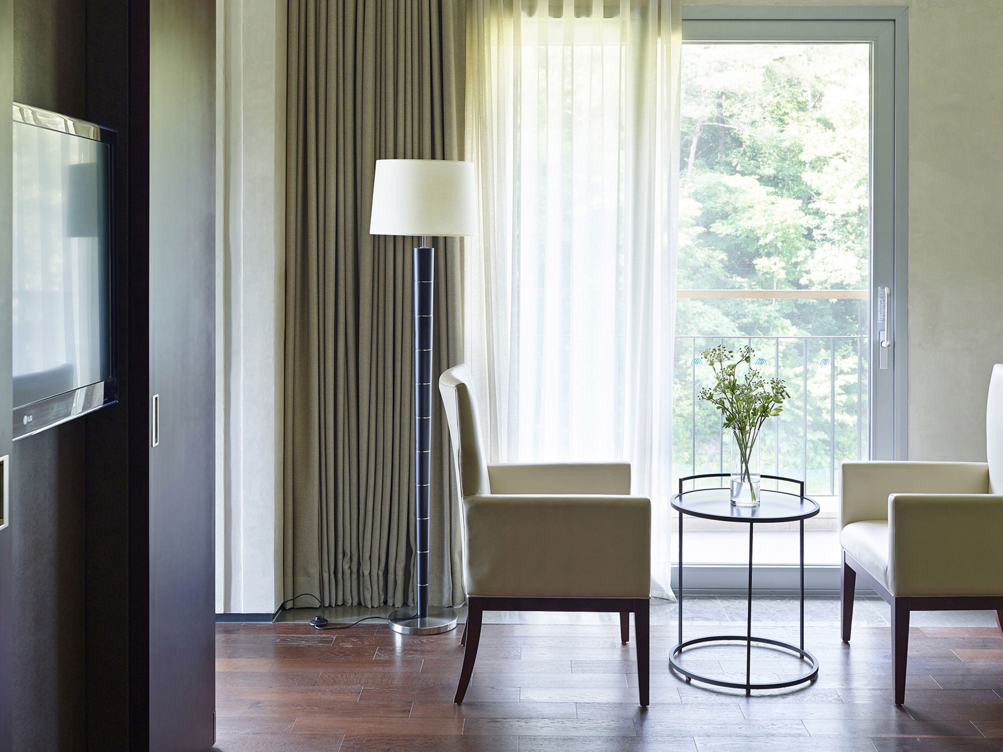 A modern hotel room with a flat-screen TV, two armchairs, a small round table with a vase of flowers, a floor lamp, and a large window with curtains.