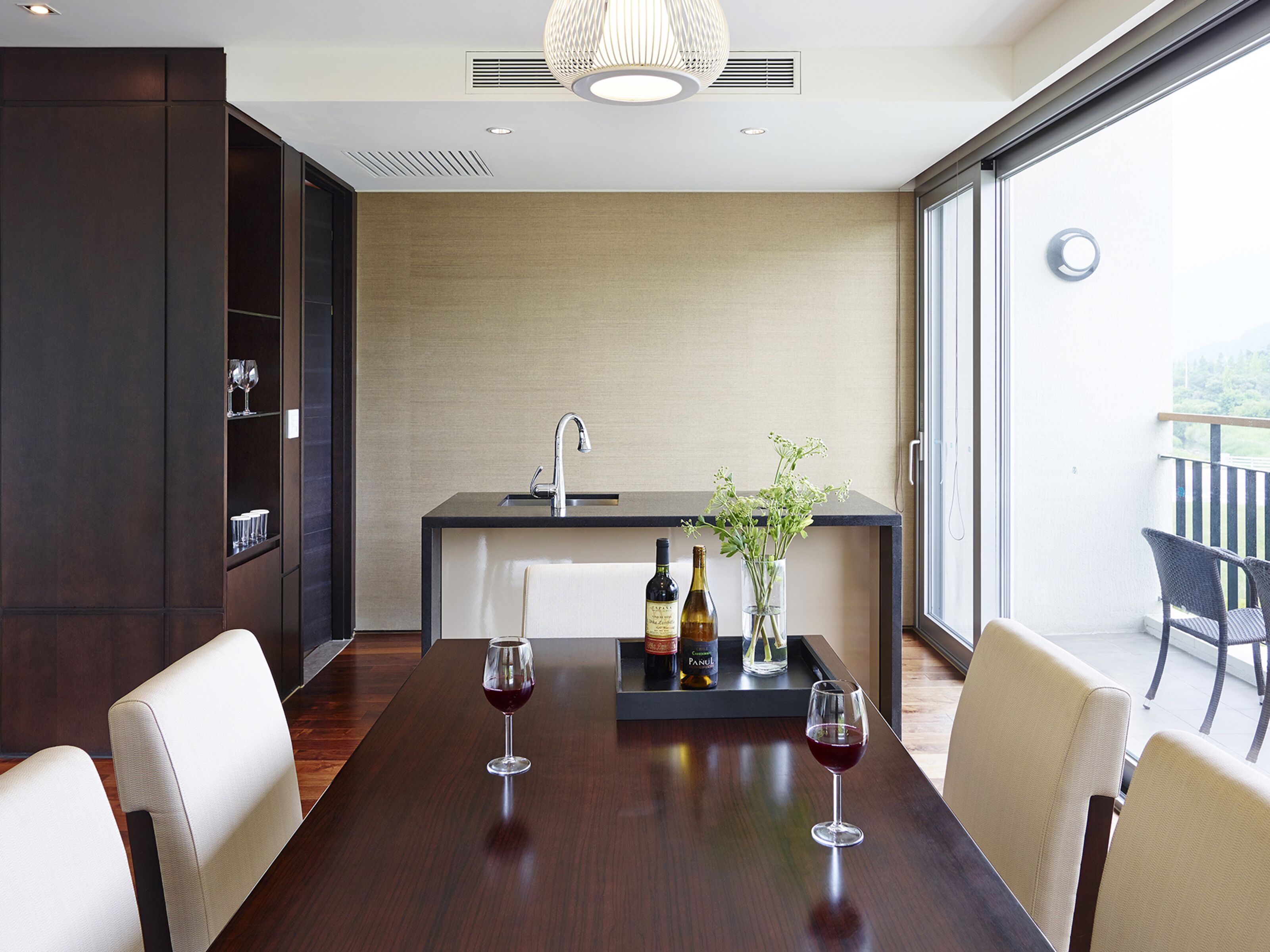A modern dining area with a dark wooden table, two wine glasses, and a bottle of wine. There is a small bar area with a sink and a vase with flowers. The room has large windows and a view of the outdoors.