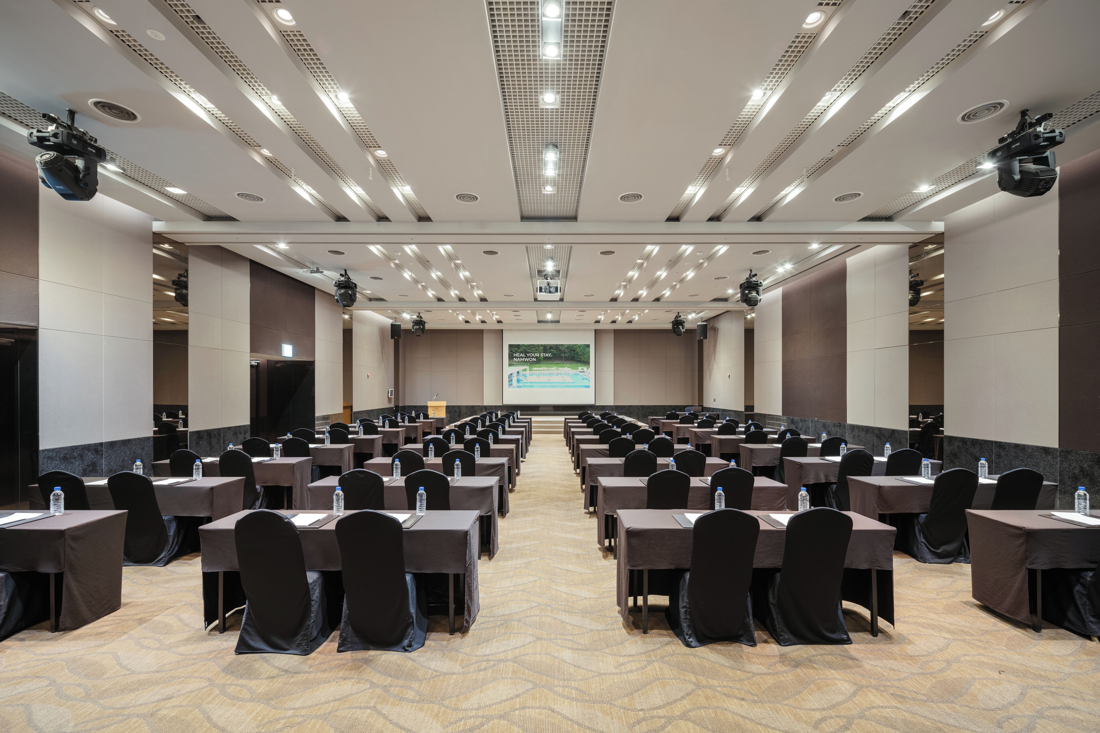 A conference room with rows of tables and chairs, a projector screen, and a carpeted floor.
