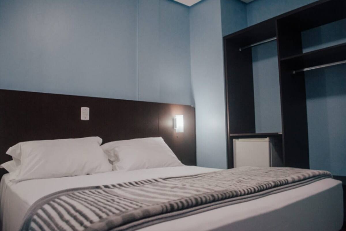 A neatly made bed with white linens and a patterned throw blanket, a dark headboard, and a wall-mounted light fixture.